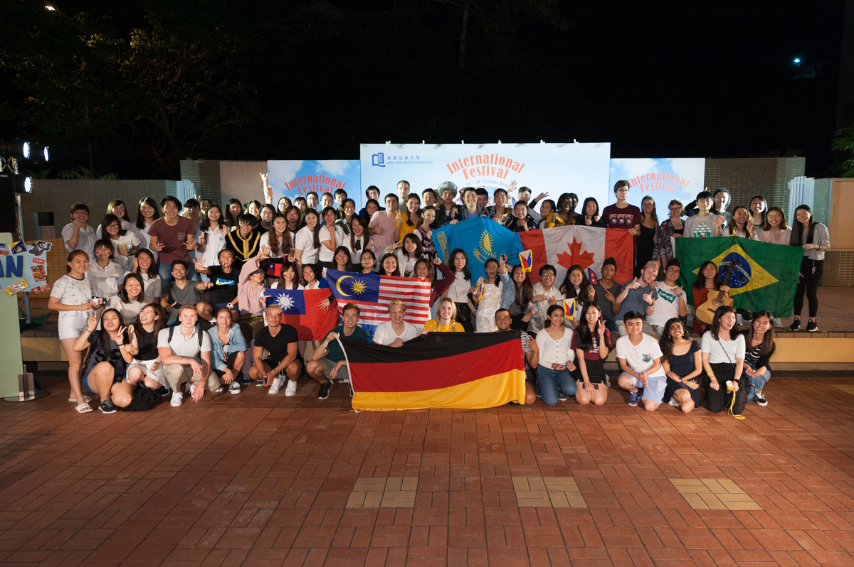 A large group of HKBU students hold up flags from their respective countries in front of the sign for the International Festival