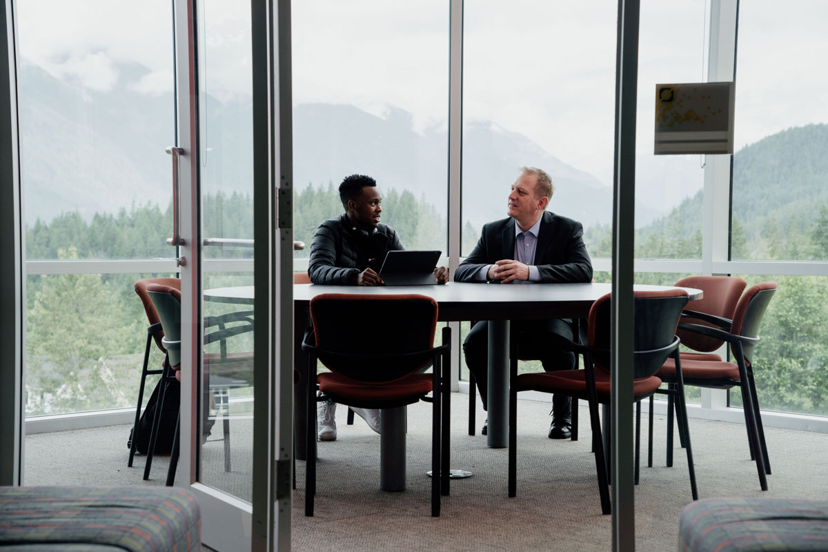 Two men sit at a conference table in a room with glass panel windows surrounded by lush forestry and mountain views